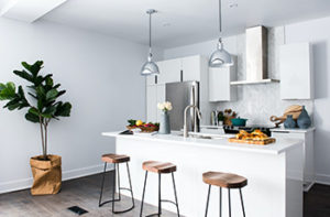 Grey-washed LVP is the flooring in this small, contemporary kitchen that features a white island, three stools, hanging light fixtures, and a stainless steel range hood.