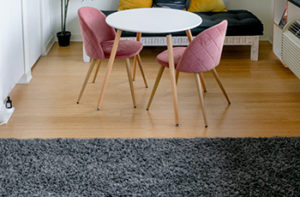 A grey microfiber area rug has been placed on a hardwood floor near a small tulip table with two pink chairs.