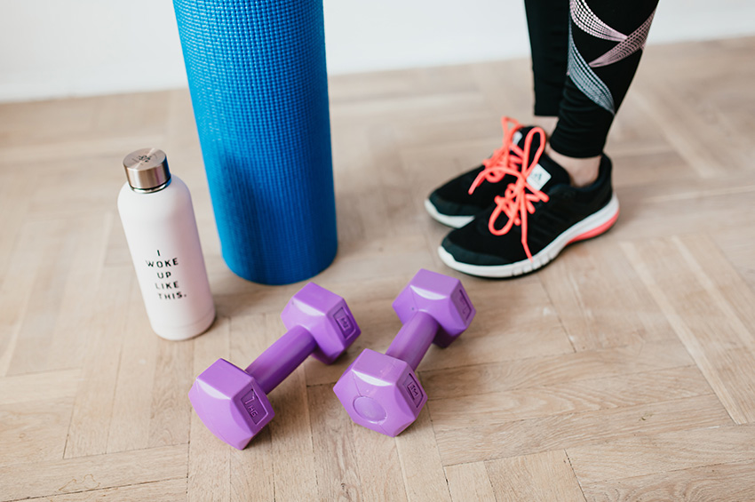 A woman in workout clothes and tennis shoes stands on light oak floors with a yoga mat, weights and pink water bottle