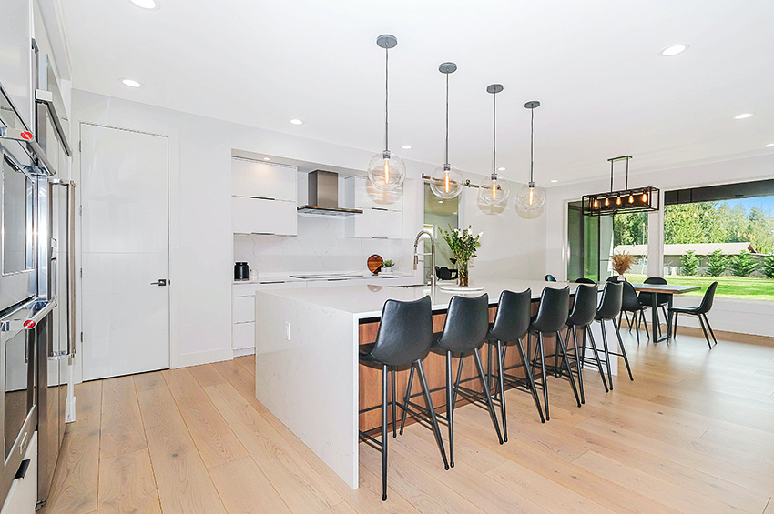 A modern kitchen features bright white walls, hanging light fixtures with glass details, a white acrylic island with six black chairs, and a large window overlooking a backyard filled with greenery.
