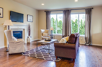 Small living room features rustic hardwood floors, white fireplace, brown area rug, couch, and floor length curtains.