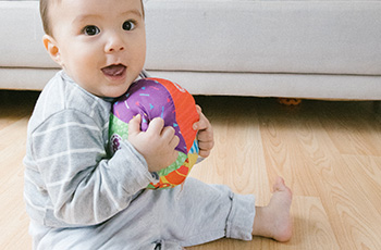 A baby sity on a hardwood floor while holding a multicolored ball and smailing.