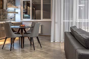 Large format tile flooring is showcased in a living and dining room area with a table, chairs, couch, and gauzy white curtains.