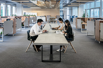 A large office space has gray commercial carpeting and a long conference table where people are working.