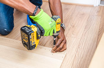 Man with drill wearing a green glove installs hardwood flooring.