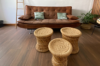 A brown couch with a contemporary look and three rattan stools are placed on hardwood flooring.