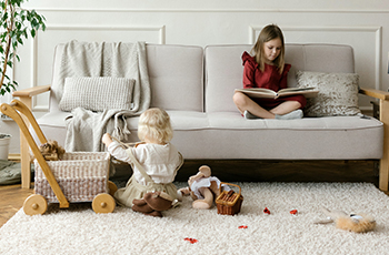 A young girl reads a book while sitting on a white couch while her baby sister plays with toys on a white area rug.