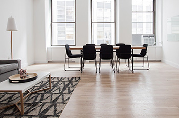 Minimalist living room design features light colored wood-look vinyl flooring, geometric rug, and dining table with eight black chairs.