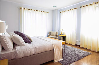 Relaxing guest bedroom showcases warm-toned hardwood floors, breezy white curtains, and a gray area rug.