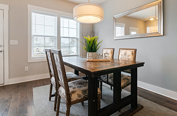 Dining room with dark wood look flooring and grey walls. There is a beige rug under the table and a plant on top of it.