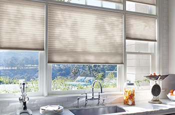 Close up of windows behind a kitchen sink. The sun is streaming through the windows that are partially covered with beige honeycomb shades.