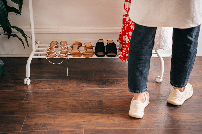 Close up shot of realistic wood look flooring in a deep color. There is a clothing rack and a person with white sneakers in the background that contrast the dark floor.