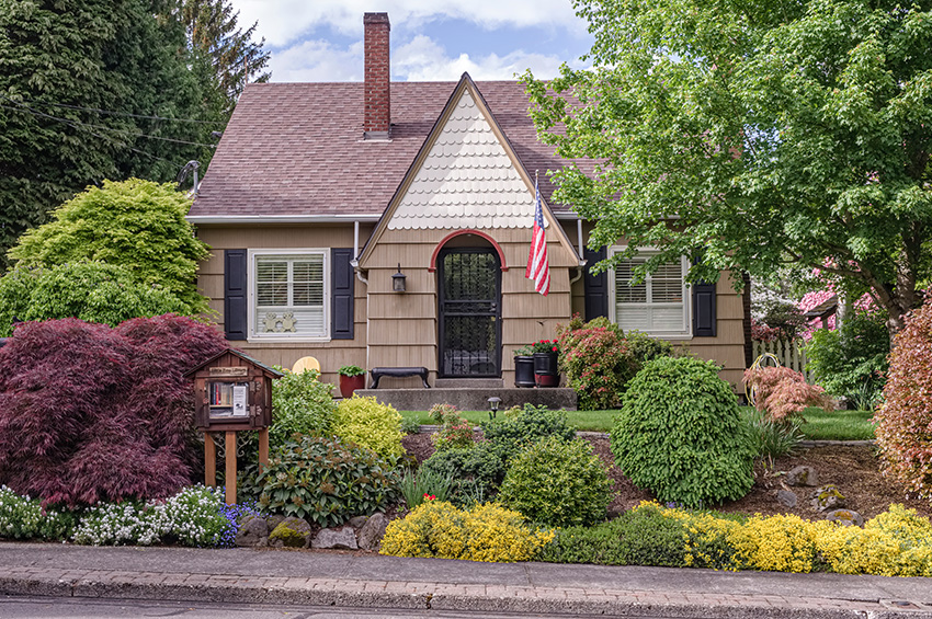 Classic Tudor home with beige siding, bushes in the front yard, and a tiny library out front.